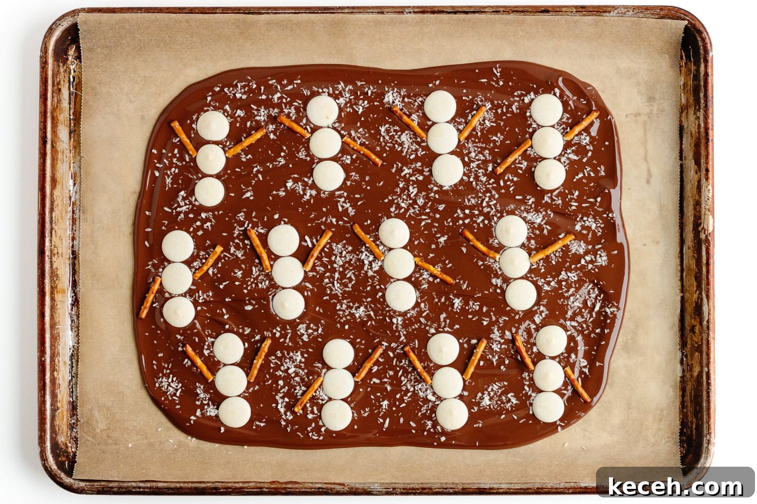 Snowman Chocolate Bark Recipe for Christmas Treats 8 Snowmen shapes being assembled on chocolate bark with three white chocolate wafers and two pretzel stick arms per snowman.